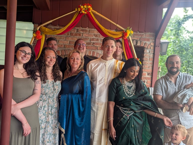 Group of eight people in colorful traditional clothing posing under red and gold draped fabric against a brick wall
