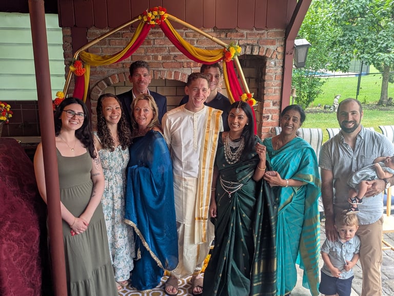 Group of people in formal attire posing together under a red and gold decorated archway at an outdoor event